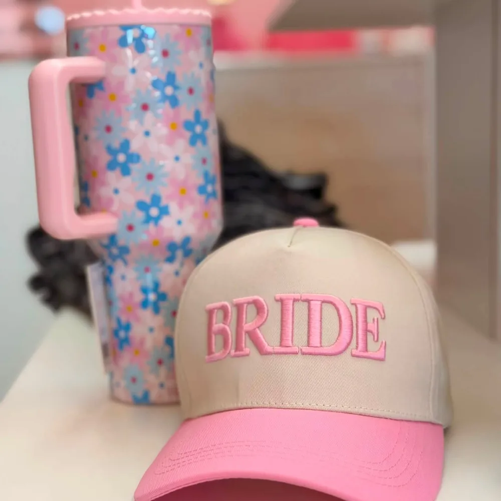 Cream trucker cap with a pink brim, embroidered BRIDE in raised pink block letters, beside a pink and blue daisy print tumbler.